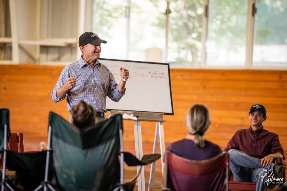 Man standing at white board speaking to a group in a stable.